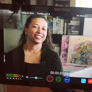 TaLisa being filmed in her studio. a close-up image of her face in the actual camera view recording screen. paintings and bookshelf blurry in the background. 